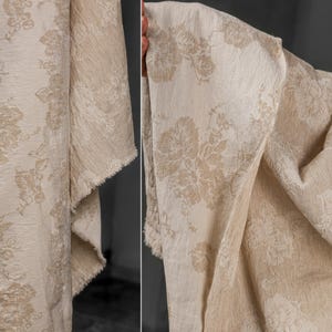 Op de afbeelding: Close-up van een beige stof met een bloemenpatroon. De stof heeft een getextureerde uitstraling en een rauwe, onafgewerkte rand. Het bloemmotief is in een lichtere tint beige, wat een subtiel contrast creëert. De stof is gedrapeerd en toont zijn textuur en ontwerp.