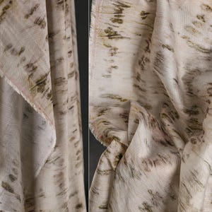 Puede incluir: Primer plano de una tela color crema con un patr&oacute;n texturizado. La tela presenta un dise&ntilde;o de pinceladas marrones y verde oliva, creando un aspecto natural y art&iacute;stico. Los bordes de la tela est&aacute;n deshilachados, lo que a&ntilde;ade encanto r&uacute;stico.