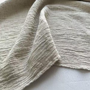 May include: Close-up of a folded, textured fabric in a neutral, light beige color. The fabric has a crinkled, woven appearance with visible horizontal lines. The edges of the fabric are slightly frayed, adding to its natural aesthetic.