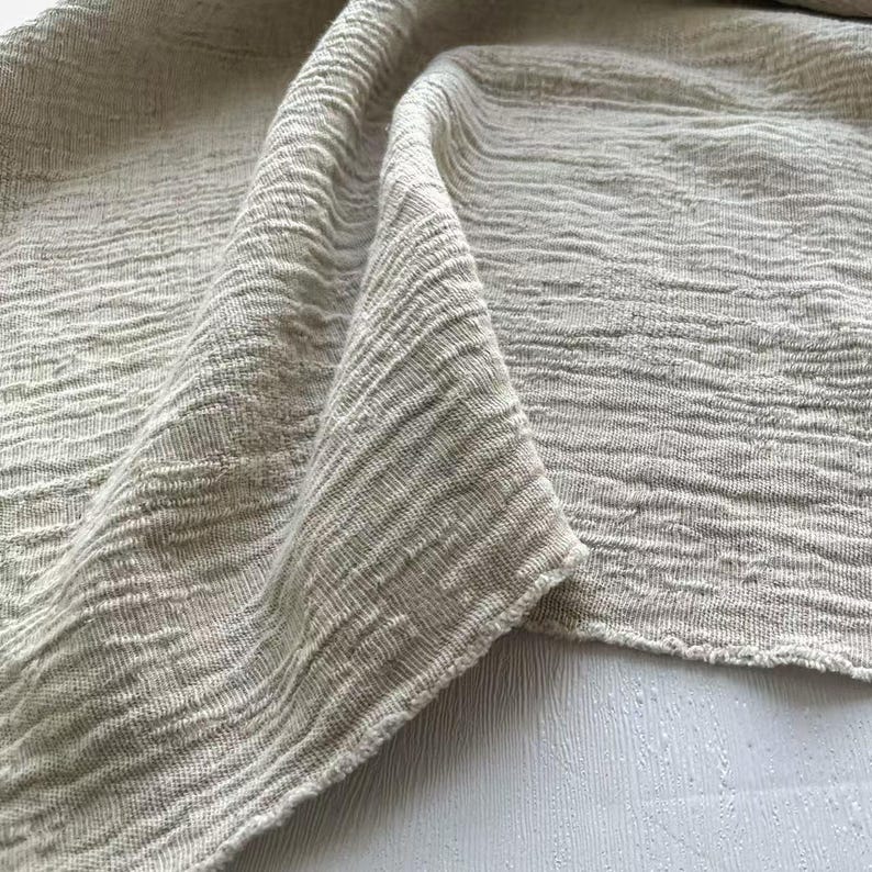 May include: Close-up of a folded, textured fabric in a neutral, light beige color. The fabric has a crinkled, woven appearance with visible horizontal lines. The edges of the fabric are slightly frayed, adding to its natural aesthetic.