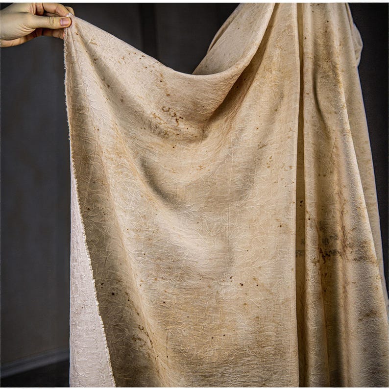 May include: A beige fabric with a textured surface and subtle patterns. The fabric has a natural, organic appearance with darker brown spots and a slightly rough texture. The material is draped, revealing its folds and the interplay of light and shadow.