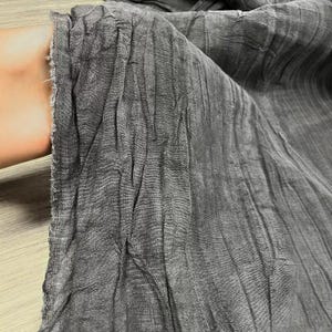 Pode incluir: Close-up de um tecido cinza com textura enrugada. O tecido parece leve e fluido, com linhas verticais e uma superfície ligeiramente irregular. A borda do tecido é visível, mostrando uma bainha crua e inacabada.