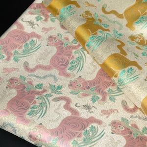 May include: Close-up of a fabric with a repeating pattern of stylized tigers in pink and gold. The tigers are interspersed with green foliage and small scorpions on a cream-colored background. The fabric is folded, showing its texture.
