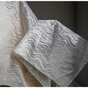 May include: Close-up of off-white fabric with a wave-like pattern. The fabric is folded, revealing layers and textures. The design is a repeating series of curved lines, creating a sense of movement and depth. The fabric appears to be soft and luxurious.