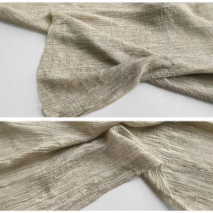May include: Close-up of beige fabric with a textured, crinkled appearance. The fabric is folded and draped, showcasing its natural, earthy tones and subtle variations in colour. The material appears to be lightweight and flowing.