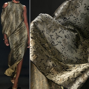 May include: A one-shoulder, olive green dress with a high neckline and a thigh-high slit. The dress is made of a fabric with a subtle pattern and a draped design. The fabric has a black script-like pattern. The dress is paired with gold-colored heels.