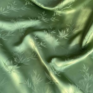 May include: Close-up of green satin fabric with a bamboo leaf pattern. The fabric has a subtle sheen and is draped in soft folds. The bamboo leaves are a lighter shade of green, creating a delicate contrast.