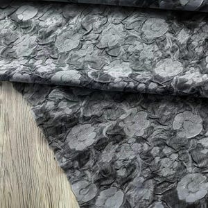 May include: Close-up of a gray fabric with a raised floral pattern. The fabric has a textured, three-dimensional appearance, with the floral design creating a layered effect. The color is a muted gray, with variations in tone.