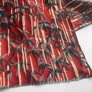 Puede incluir: Tela roja con un estampado de bambú. El diseño presenta tallos de bambú blancos y hojas de bambú verdes y grises. La tela tiene un aspecto texturizado y un borde deshilachado.
