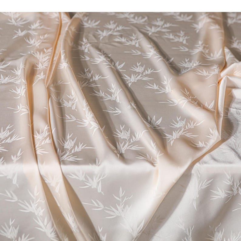 May include: A close-up of a beige fabric with a white bamboo leaf pattern. The fabric is draped, creating soft folds and shadows. The material has a subtle sheen, suggesting a silk or satin texture. The overall effect is elegant and natural.