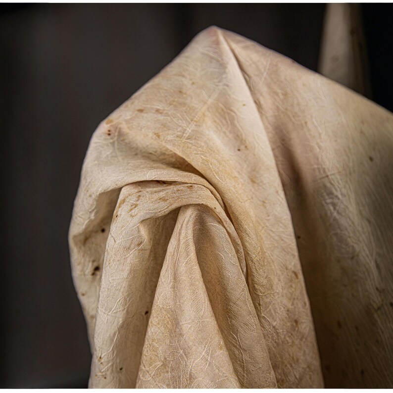 May include: Close-up of a beige fabric with a textured surface and subtle brown spots. The fabric is draped, creating folds and shadows. The material appears to be natural, possibly linen or a similar textile.