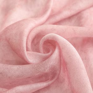 May include: Close-up of a soft, flowing fabric in a delicate pink hue. The material is finely textured, with subtle variations in color and light, creating a gentle, ethereal effect. The fabric is draped in graceful folds and swirls, suggesting a lightweight textile.
