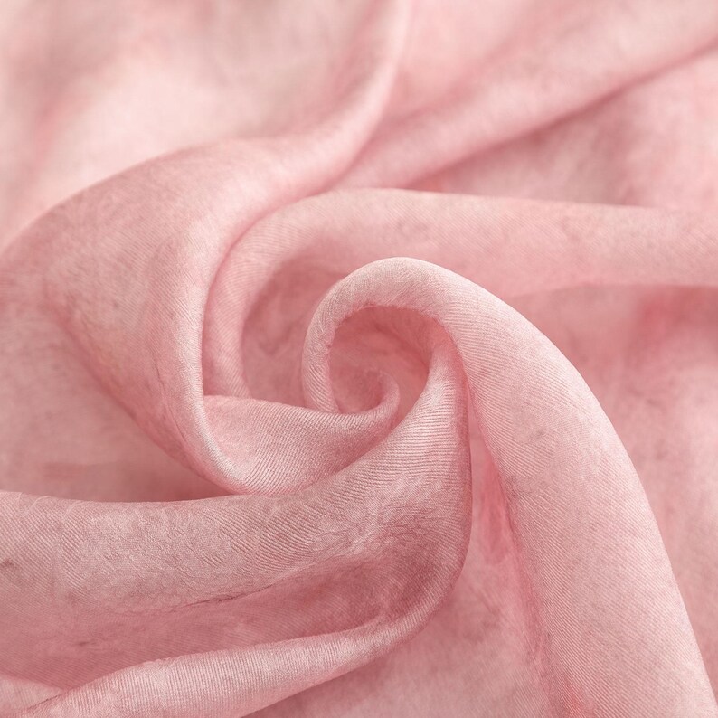 May include: Close-up of a soft, flowing fabric in a delicate pink hue. The material is finely textured, with subtle variations in color and light, creating a gentle, ethereal effect. The fabric is draped in graceful folds and swirls, suggesting a lightweight textile.