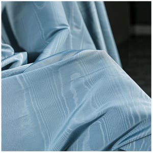 May include: Close-up of a light blue fabric with a subtle, textured pattern. The fabric is draped, showing folds and shadows that highlight its soft texture. The material appears to be lightweight and flowing, suitable for drapery or apparel.