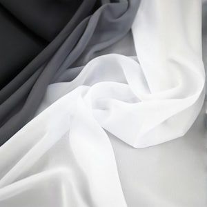 May include: A close-up of a flowing fabric with a gradient effect. The fabric transitions from black to gray to white, creating a soft, ethereal look. The material appears lightweight and sheer, with gentle folds and curves.