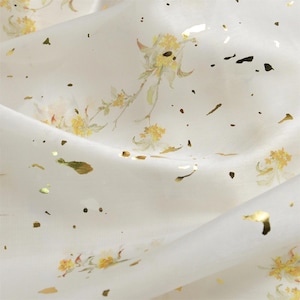 May include: Close-up of a white fabric with a delicate floral pattern in yellow and green. Gold foil accents are scattered across the surface. The fabric has a soft, flowing appearance, suggesting it is lightweight.