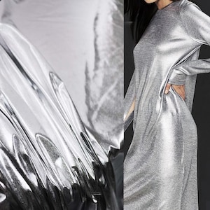 Pode incluir: Um vestido prateado de manga comprida com uma silhueta fluida. O tecido tem um brilho metálico, refletindo a luz e criando um efeito cintilante. O vestido apresenta um decote alto e uma fenda lateral.