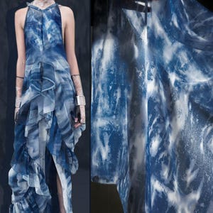 May include: A flowing, sleeveless dress in shades of blue and white, with a watercolour-like pattern. The dress has a halter neckline and layered ruffles, creating a dynamic silhouette. The fabric appears lightweight and airy, with a high slit.