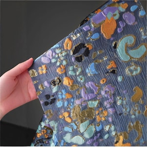 May include: Close-up of a textured fabric with a dark blue background. The fabric features a pattern of colorful, abstract shapes in shades of orange, purple, blue, and gold. The fabric is being held by a hand.