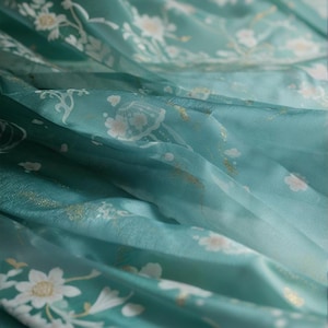 May include: Close-up of a flowing, sheer fabric in shades of teal and turquoise, adorned with delicate white floral patterns and subtle gold accents. The fabric's texture is soft and flowing, suggesting a lightweight material like chiffon or silk.