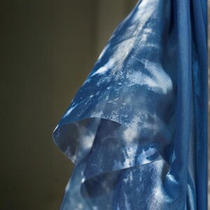 May include: Close-up of a flowing, sheer fabric in shades of blue with a white, cloud-like pattern. The fabric is draped, revealing layers and textures. The overall effect is soft and airy, suggesting a scarf or shawl.