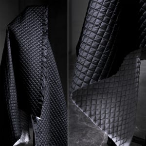 May include: Black quilted fabric with a diamond pattern. The fabric is draped over a surface, showcasing its texture and design. The reverse side of the fabric is a lighter gray color, also with a diamond pattern.