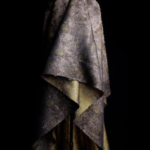 May include: A piece of fabric featuring a dark, distressed charcoal and brown exterior with a metallic gold interior. The material has a textured, weathered appearance with visible folds and drapes, showcasing the contrast between the dark outer surface and the bright, reflective gold lining.