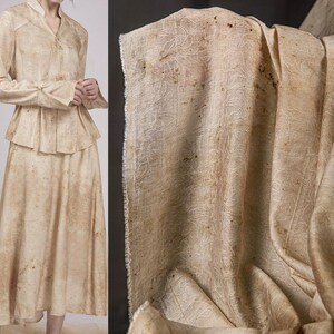 May include: A two-piece beige outfit with a jacket and skirt, alongside a close-up of matching fabric. The fabric has a subtle, organic pattern with brown speckles. The jacket features a peplum design and long sleeves. The skirt is a midi length.