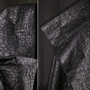 Könnte beinhalten: Nahaufnahme eines schwarzen Kleidungsstücks mit Kapuze und einem strukturierten, krokodilartigen Muster. Der Stoff scheint Leder oder ein lederähnliches Material zu sein. Die Kapuze ist umgeklappt und zeigt das Innenfutter.