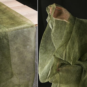 May include: A sheer, olive green fabric draped over a wooden surface and held up by a hand. The fabric has a textured appearance and a subtle, organic pattern. The overall aesthetic is soft and ethereal, suggesting a decorative textile.