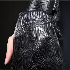 Puede incluir: Primer plano de una tela negra tejida con un patrón de tejido de cesta. La tela está drapeada, mostrando la interacción de la luz y la sombra en su superficie. El material parece flexible y posiblemente se utilice para ropa o tapicería.