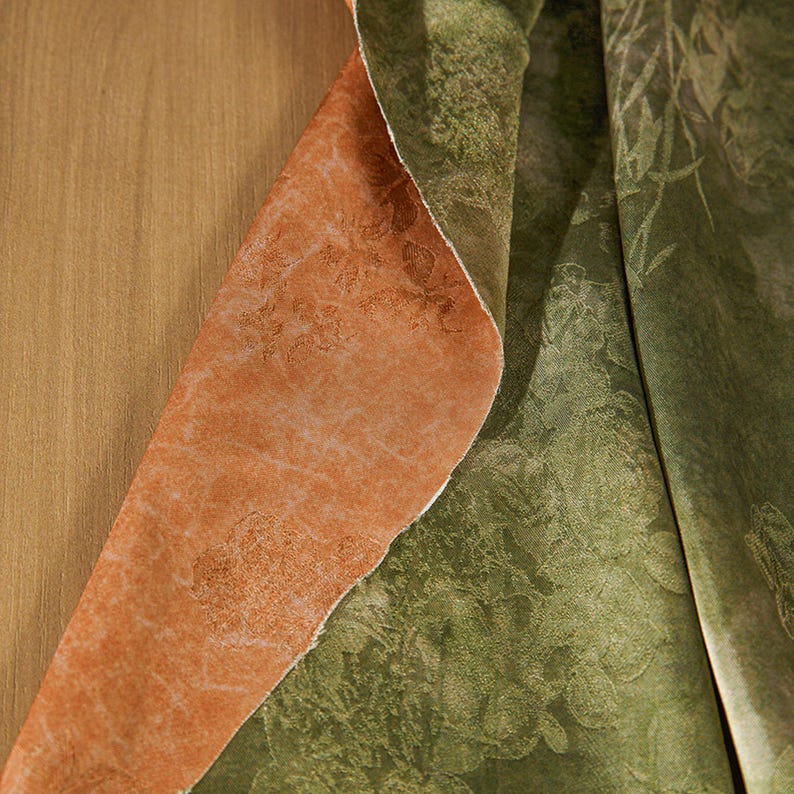 May include: Close-up of two fabric samples. One is a burnt orange color with a textured surface and a floral pattern. The other is a green fabric with a floral design. The fabrics are layered, revealing their contrasting colors and textures.