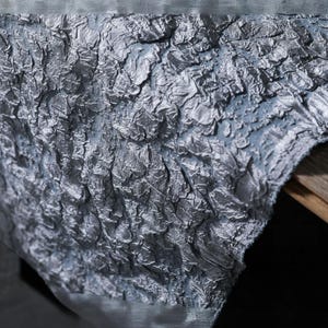 Op de afbeelding: Close-up van een getextureerde, zilvergrijze stof met een gekreukeld uiterlijk. De stof heeft een gelaagd, driedimensionaal effect, met subtiele variaties in toon. Het materiaal lijkt over een houten oppervlak te zijn gedrapeerd, met een vleugje donkerdere achtergrond.