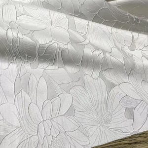Könnte beinhalten: Nahaufnahme eines weißen Stoffes mit floralem Muster. Das Design zeigt geprägte Blumen und Blätter, wodurch eine strukturierte Oberfläche entsteht. Der Stoff hat einen dezenten Glanz, und das Muster ist gleichmäßig.