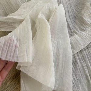 May include: Close-up of white fabric with a textured, pleated design. The fabric is draped and folded, creating shadows and highlights. The material appears lightweight and semi-transparent, suggesting a delicate textile.