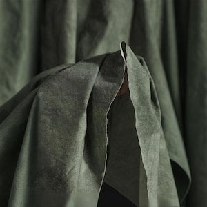 Könnte beinhalten: Nahaufnahme eines olivgrünen Stoffes mit strukturierter Oberfläche. Der Stoff ist gefaltet und zeigt eine hellere Unterseite. Die Falten erzeugen Schatten und Tiefe und heben die Textur des Materials hervor. Die Gesamtästhetik ist weich und gedämpft.