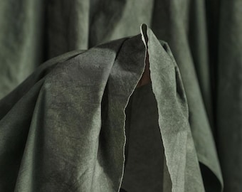 Puede incluir: Primer plano de tela verde oliva con superficie texturizada. La tela está doblada, revelando un reverso más claro. Los pliegues crean sombras y profundidad, resaltando la textura del material. La estética general es suave y apagada.