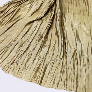 May include: Close-up of a flowing, textured fabric in a golden-olive colour. The material is heavily draped, with vertical and diagonal folds creating a dynamic pattern. The fabric has a subtle sheen, suggesting a silk or satin-like quality.