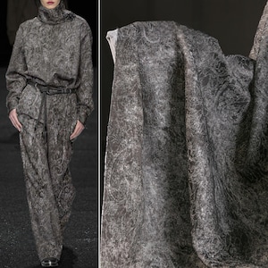 Puede incluir: Un conjunto de dos piezas y una muestra de tela con un estampado de cachemira gris oscuro. El conjunto incluye un top de manga larga con cuello alto y pantalones de pernera ancha, ceñidos con un cinturón. La muestra de tela muestra el mismo diseño intrincado.