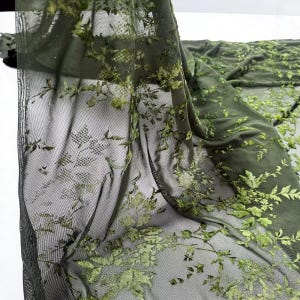 May include: Olive green sheer fabric with embroidered floral and leaf patterns. The delicate design features raised, textured details in a lighter shade of green, creating a layered effect. The fabric is draped, showcasing its flowing quality and intricate design.