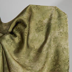 May include: A close-up of a draped fabric in shades of olive green. The fabric features a subtle floral pattern, with visible details of leaves and flowers. The texture appears soft and slightly textured, with variations in light and shadow.