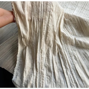 Op de afbeelding: Close-up van een beige, geplooide stof met verticale lijnen en een getextureerd uiterlijk. De stof wordt door een hand vastgehouden, waardoor de rafelige rand van het materiaal zichtbaar wordt. De stof heeft een natuurlijke, licht gekreukelde uitstraling.