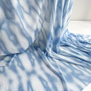 May include: A roll of blue and white tie-dye fabric. The fabric is a soft, flowing material with a textured, dotted pattern. The blue dye creates a marbled effect across the white background. The fabric is likely used for clothing or home decor.