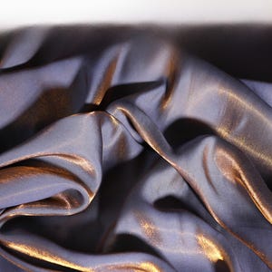 Könnte beinhalten: Nahaufnahme eines drapierten Stoffes mit schimmerndem, irisierendem Effekt. Der Stoff ist hauptsächlich in einem gedämpften Lila-Grau gehalten, mit goldenen Glanzlichtern, die das Licht einfangen. Die Falten und Texturen erzeugen ein Gefühl von Tiefe und Bewegung.