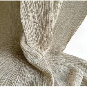 May include: A close-up shot of a light beige fabric with a textured, ribbed surface. The fabric is draped, forming folds and shadows that accentuate its texture and the way light interacts with it. The material looks soft and flowing.