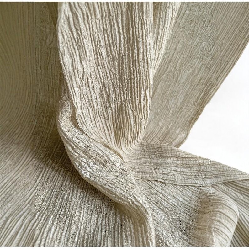 May include: A close-up shot of a light beige fabric with a textured, ribbed surface. The fabric is draped, forming folds and shadows that accentuate its texture and the way light interacts with it. The material looks soft and flowing.