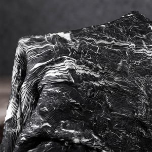 Może przedstawiać: Kawałek tkaniny o kontrastującym czarno-białym wzorze z gęstymi, falistymi i warstwowymi liniami, które tworzą topograficzny lub geologiczny wygląd. Materiał ma głęboko pomarszczoną, plisowaną teksturę na całej swojej powierzchni.