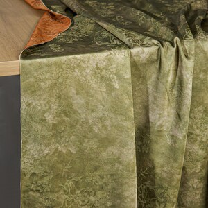 May include: A draped fabric with a forest-like pattern in shades of green and beige. The fabric has a textured appearance, with detailed botanical designs. The reverse side of the fabric is a solid burnt orange color, visible where the fabric is folded.