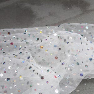May include: Close-up of white tulle fabric scattered with iridescent sequins. The sequins are various sizes and colors, including silver, blue, pink, and gold. The fabric is slightly wrinkled, creating shadows and depth.