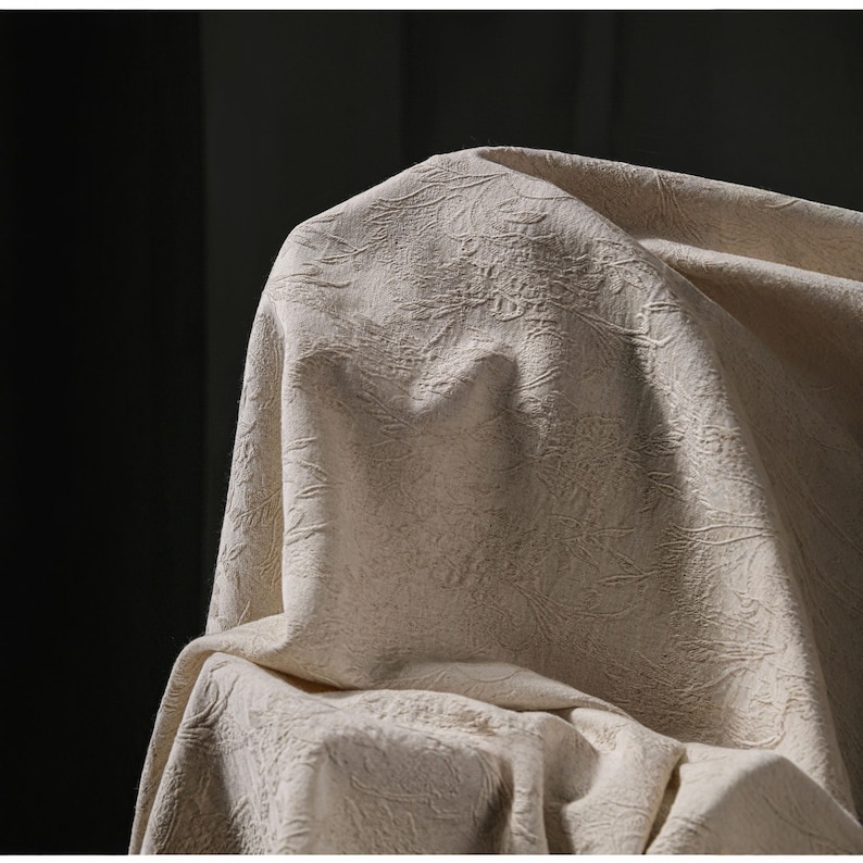 May include: Close-up of a beige fabric with a textured, floral-like pattern. The fabric is draped, creating soft folds and shadows. The material appears to be thick and possibly used for upholstery or drapery.